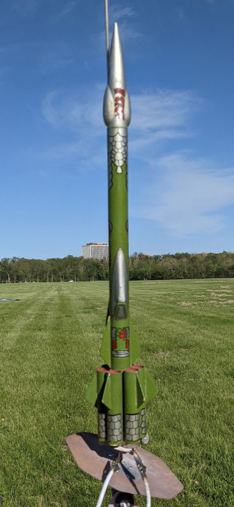 An Estes Torellian Invader model from the late 1970s / early 1980s. This is a green and silver tube-fin rocket with science-fiction details. The rocket is on a launch pad on a sunny day in the park.